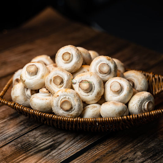 A basket of white button mushrooms sits on a wooden surface. The mushrooms are all different sizes and shapes, and they have a smooth, white skin.