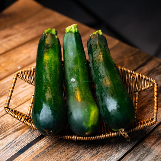 Three green zucchinis sit on a wooden basket. The zucchinis are long and cylindrical, with smooth, dark green skin.