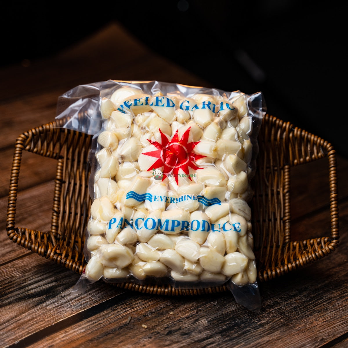 A packet of peeled garlic cloves sits on a wooden basket. The cloves are white. They are packaged in a clear plastic bag with a red label.