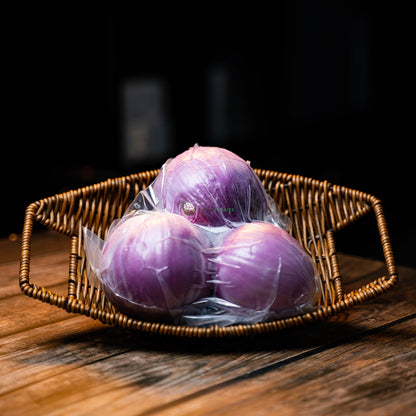 Three peeled Bombay onions in plastic wrap, resting on a wooden surface inside a basket.