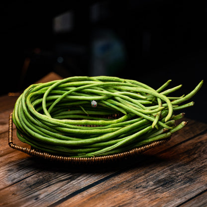 A bunch of green long beans, laid on a wicker basket on a wooden surface. The beans are fresh and slender, with a slightly curved shape.