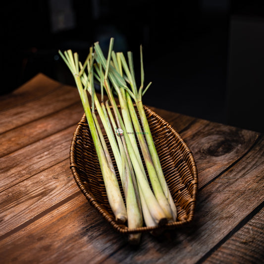 A bunch of lemongrass, with tall, green stalks and a strong, citrusy aroma, sits in a basket on a wooden surface.