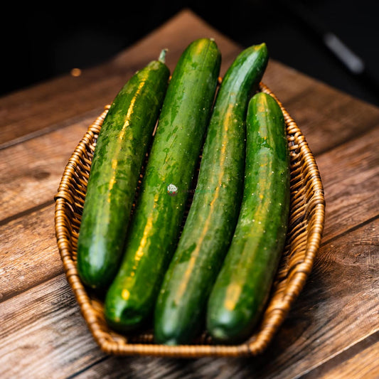 Four Japanese cucumbers, green and firm, sit in a basket on a wooden surface. They have thin, smooth skin and a slightly sweet flavor.